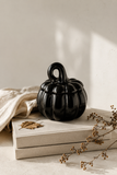 A glossy black pumpkin-shaped decorative container with a lid and loop handle, styled on top of two neutral-toned books with a beige cloth and dried botanicals in a softly lit indoor setting.